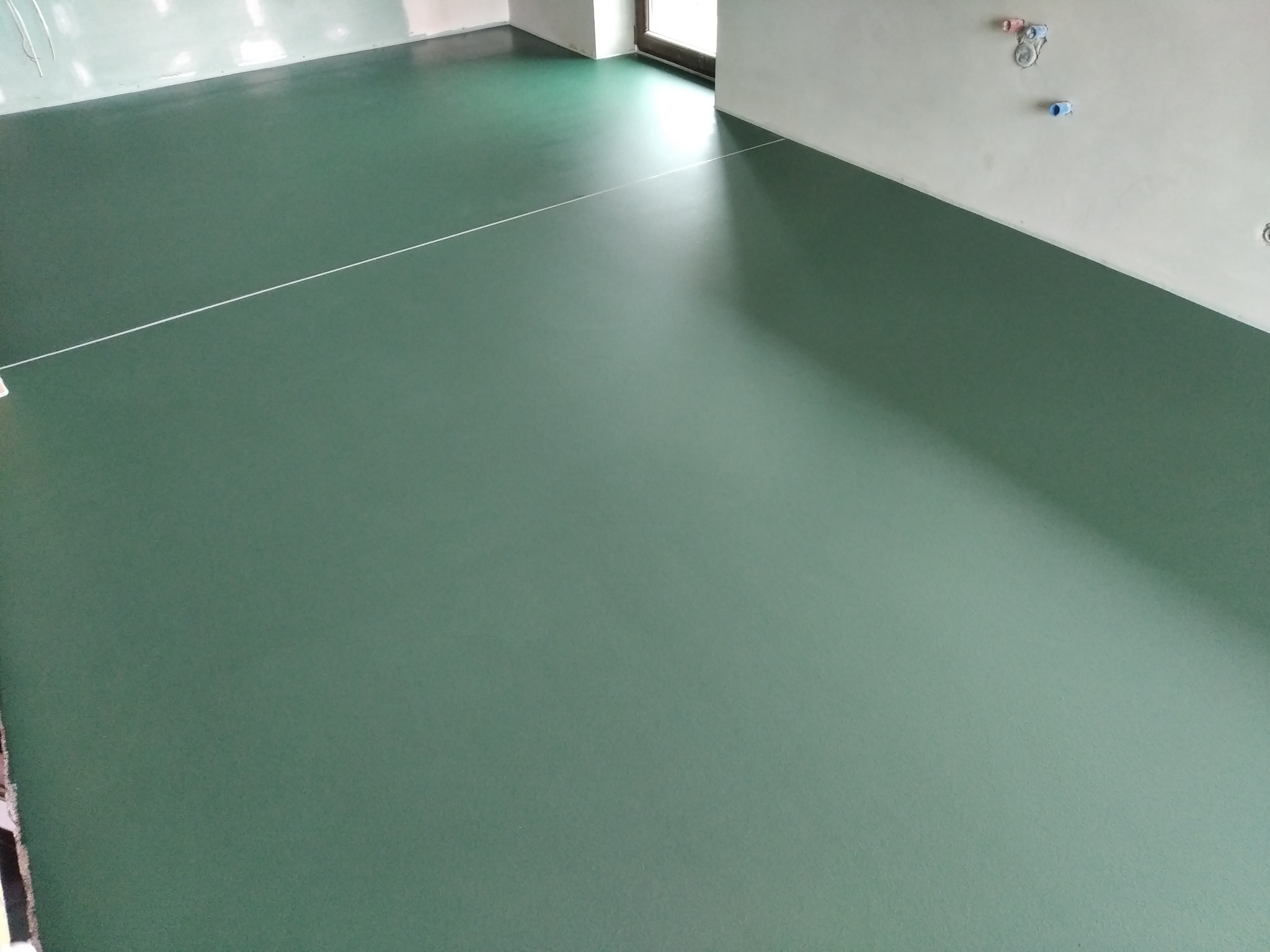 Świeżo wylana, jednolita, ciemnozielona podłoga w pokoju z białymi ścianami i oknem, widoczna cienka, biała linka wyznaczająca linię prostą.