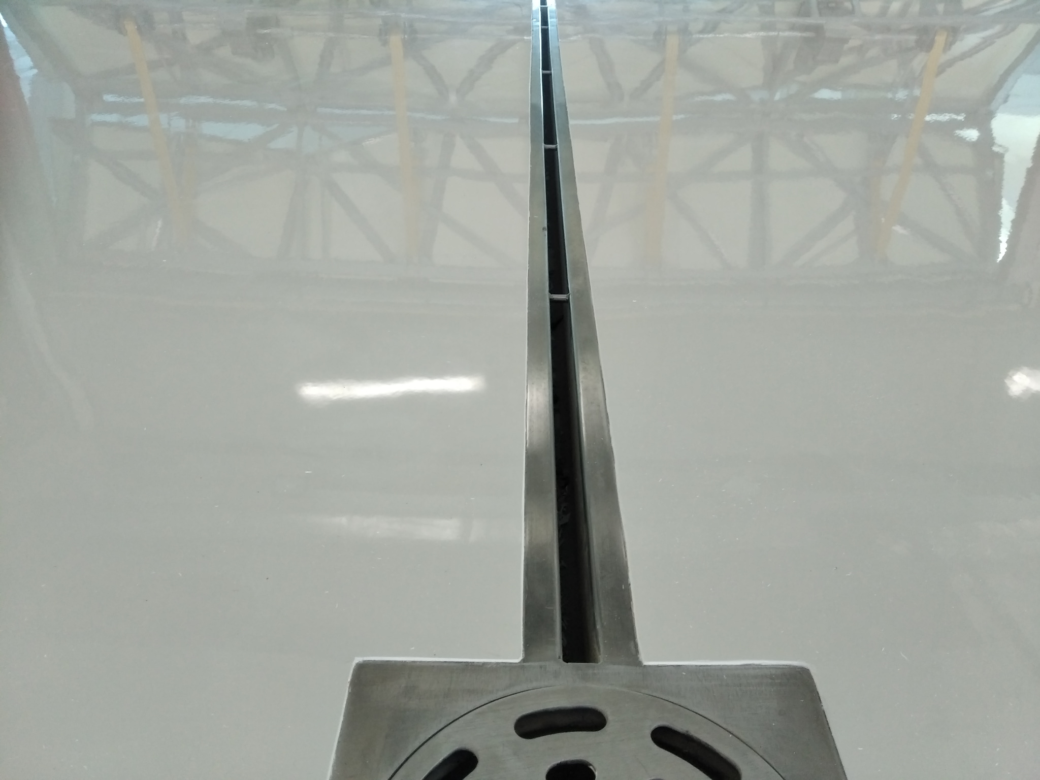 Szara, gładka powierzchnia podłogi z odpływem liniowym ze stali nierdzewnej, widoczne odbicie konstrukcji dachu.