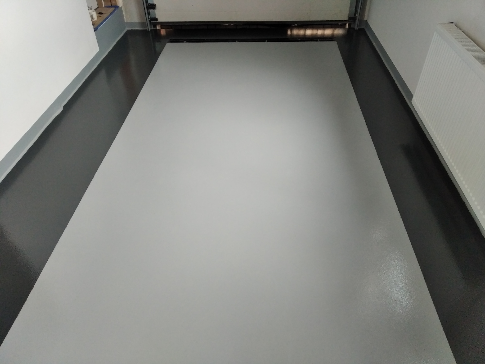 Gładka, jasnoszara podłoga w garażu z widoczną bramą garażową i grzejnikiem na ścianie.