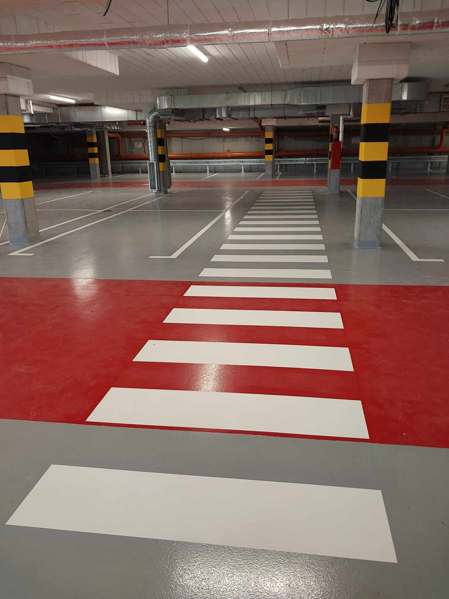 Oznakowanie poziome w garażu podziemnym: czerwona strefa z białymi pasami przejścia dla pieszych na szarej posadzce, słupy z czarno-żółtymi pasami, widoczne instalacje pod sufitem.
