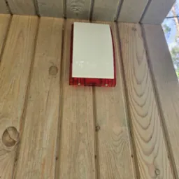 Zewnętrzna syrena alarmowa z białą obudową i czerwoną podstawą, zamontowana na drewnianej ścianie z widocznymi sękami.