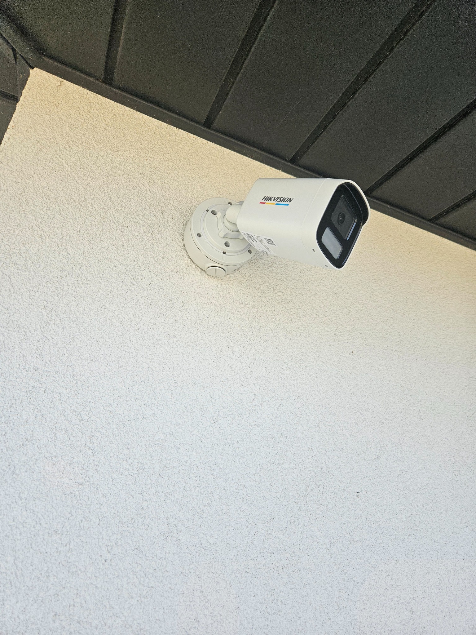Biała kamera przemysłowa marki Hikvision zamontowana na jasnej, chropowatej ścianie budynku pod ciemnym okapem.