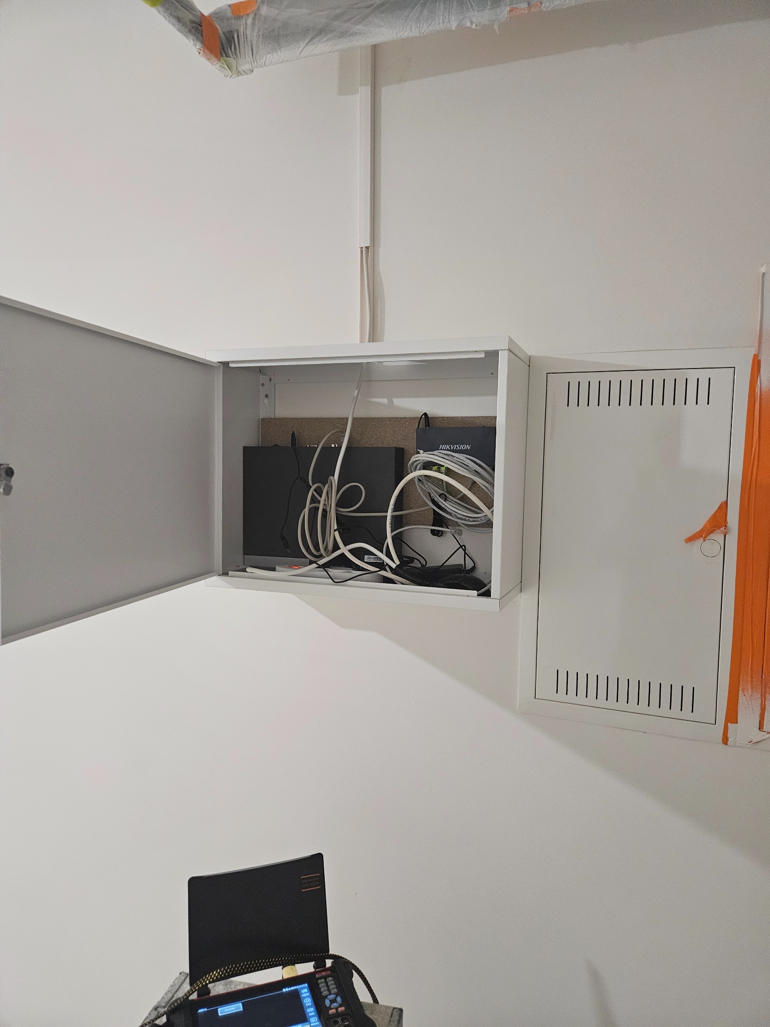 Metalowa szafka z otwartymi drzwiczkami, wewnątrz router Hikvision i splątane kable, obok druga szafka z wentylacją, widoczny tester sieci na dole kadru, białe ściany.
