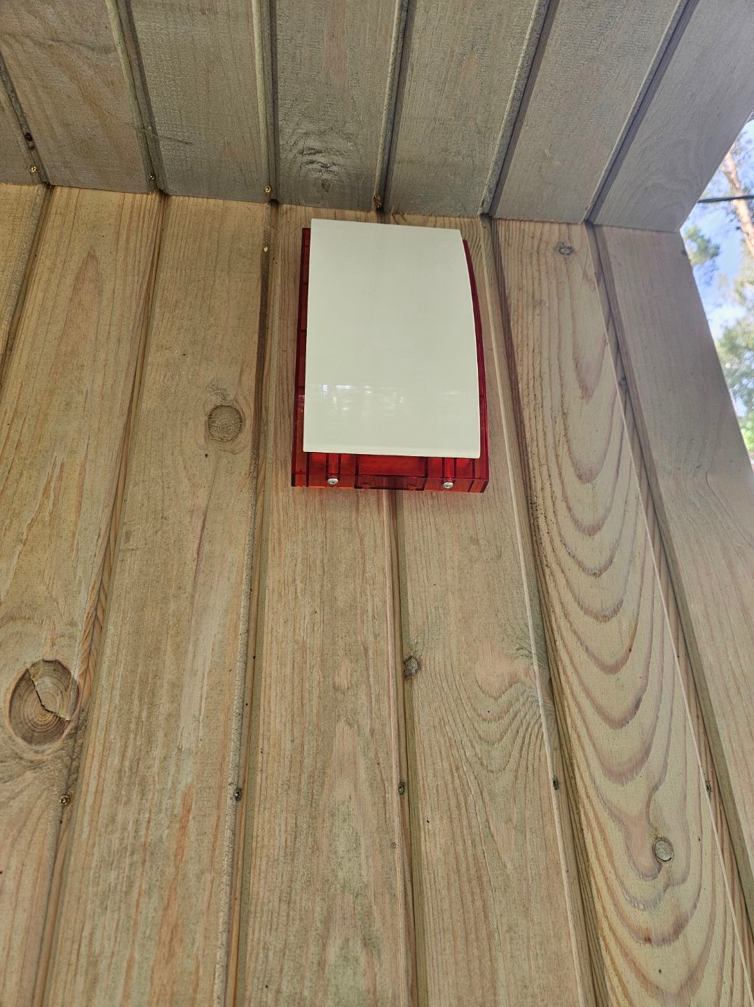 Zewnętrzna syrena alarmowa z białą obudową i czerwoną podstawą, zamontowana na drewnianej ścianie z widocznymi sękami.