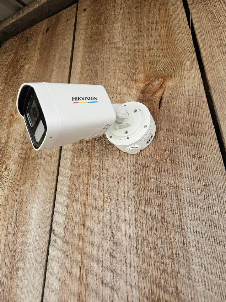 Biała kamera monitoringu Hikvision zamontowana na pionowej drewnianej ścianie z widocznym logo producenta.