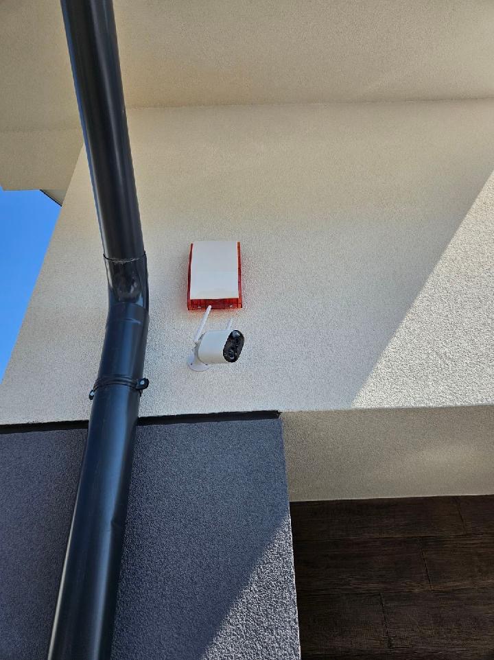 Zewnętrzna kamera monitoringu z antenami i sygnalizator alarmowy zamontowane na elewacji budynku, widoczny fragment rury spustowej.