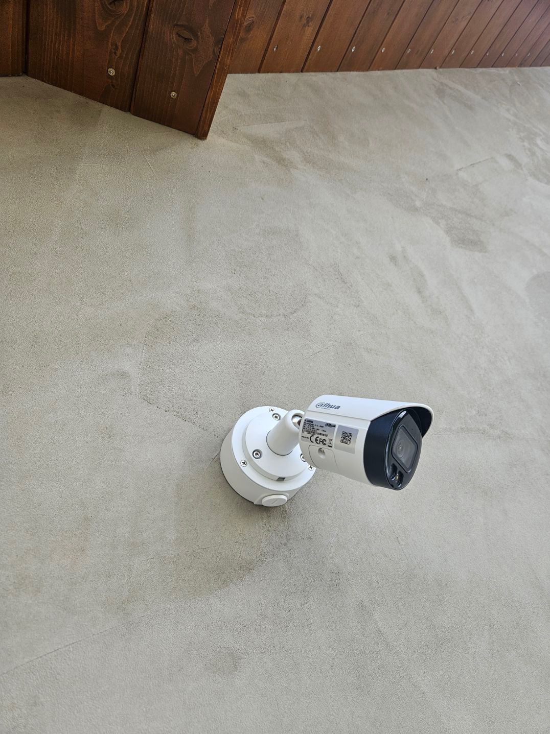 Biała kamera monitoringu Dahua zamontowana na szarej ścianie zewnętrznej budynku, widoczny fragment drewnianego okapu dachu w górnym rogu.