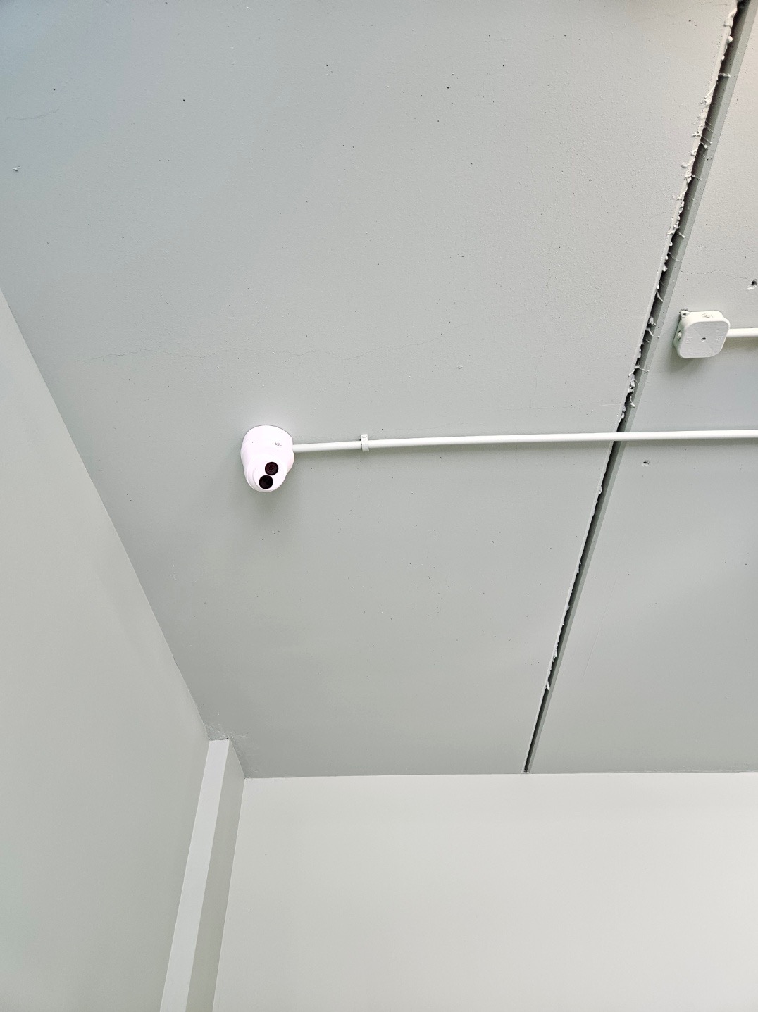 Biała kamera monitoringu kopułkowa zamontowana na szarym suficie podwieszanym z widocznymi łączeniami płyt i instalacją elektryczną w białej rurce.