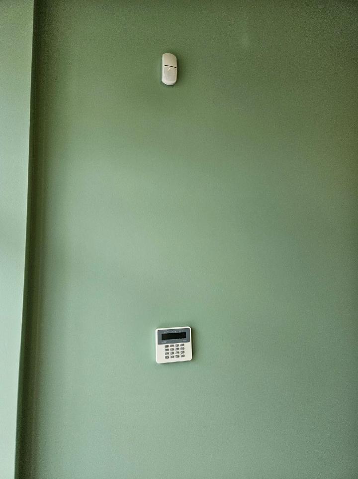 Biały panel sterowania alarmem z wyświetlaczem i klawiaturą numeryczną, zamontowany na zielonej ścianie, z czujnikiem ruchu powyżej.