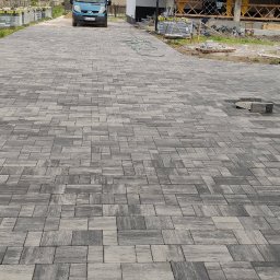Kam-Bud usługi remontowo-budowlane - Ułożona kostka brukowa w odcieniach szarości na podjeździe, widoczny fragment niezakończonej pracy z brakiem kilku kostek, w tle samochód i elementy budowy.