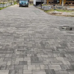 Ułożona kostka brukowa w odcieniach szarości na podjeździe, widoczny fragment niezakończonej pracy z brakiem kilku kostek, w tle samochód i elementy budowy.
