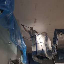 Kam-Bud usługi remontowo-budowlane - Pracownik w kombinezonie ochronnym maluje sufit agregatem malarskim, zabezpieczony folią budowlaną, widoczny regał metalowy i brama garażowa.