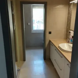 Nowoczesna łazienka z beżowymi płytkami, białymi szafkami pod umywalką z drewnianym blatem i widokiem na okno w drugim pomieszczeniu, uchwycona z perspektywy korytarza podczas remontu.