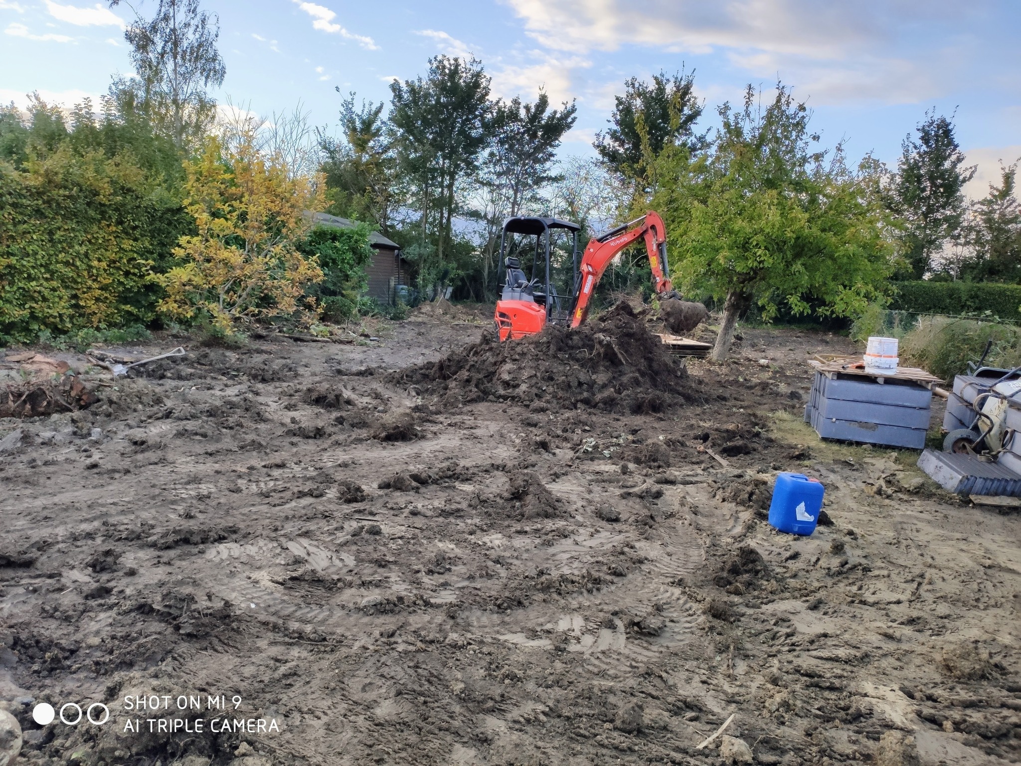 Niewielka, pomarańczowa koparka podczas prac ziemnych na grząskim, błotnistym terenie z drzewami w tle.