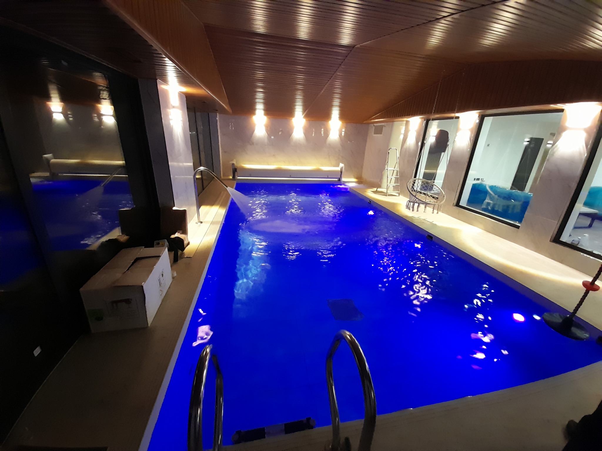 Kryty basen z niebieskim oświetleniem, widok z perspektywy schodów wejściowych, drewniany sufit i duże okna.