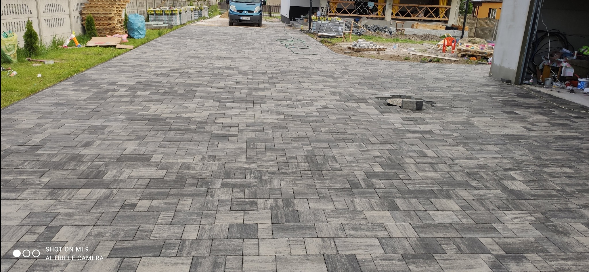Ułożona kostka brukowa w odcieniach szarości na podjeździe, widoczny fragment niezakończonej pracy z brakiem kilku kostek, w tle samochód i elementy budowy.