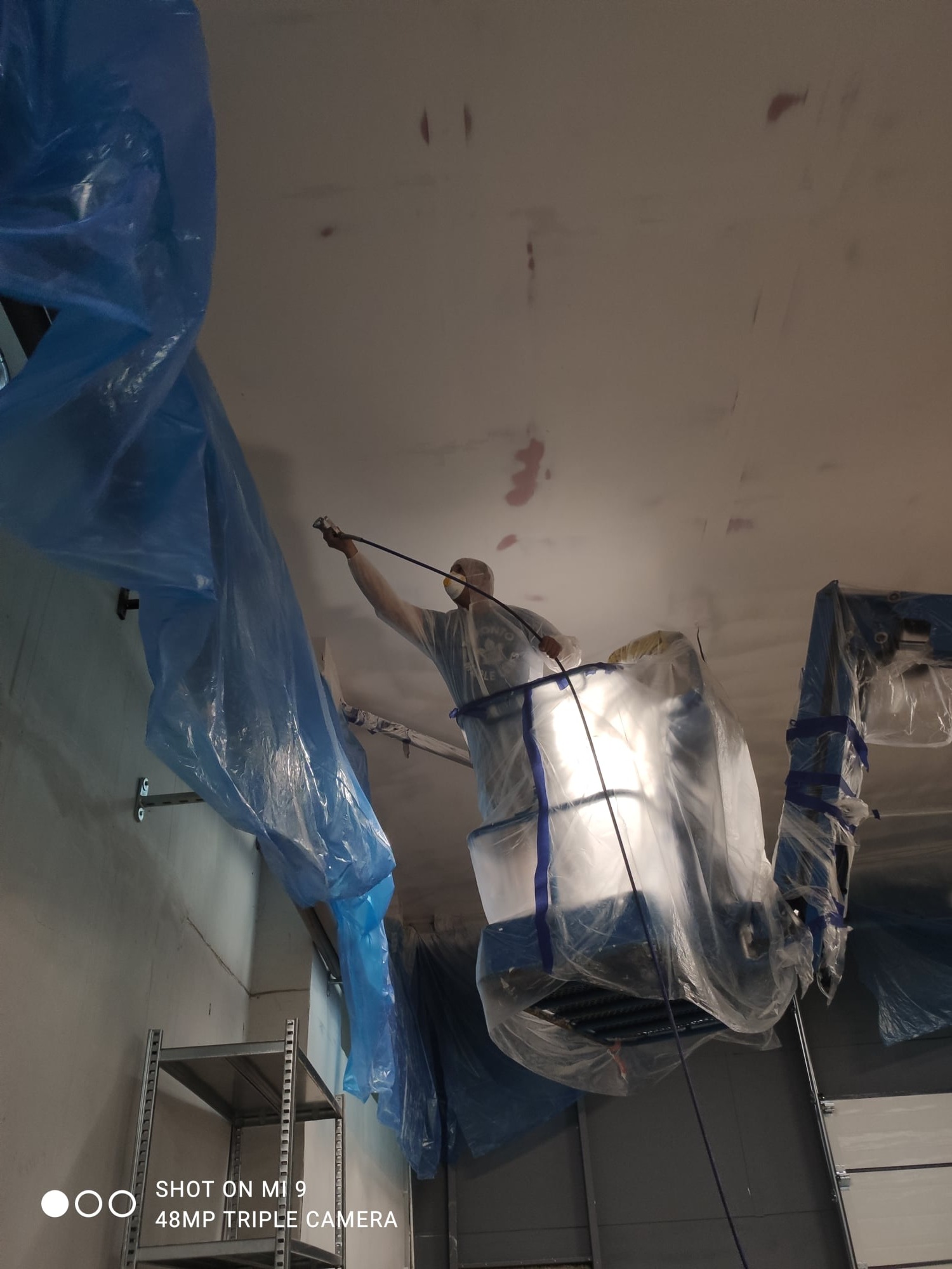 Pracownik w kombinezonie ochronnym maluje sufit agregatem malarskim, zabezpieczony folią budowlaną, widoczny regał metalowy i brama garażowa.