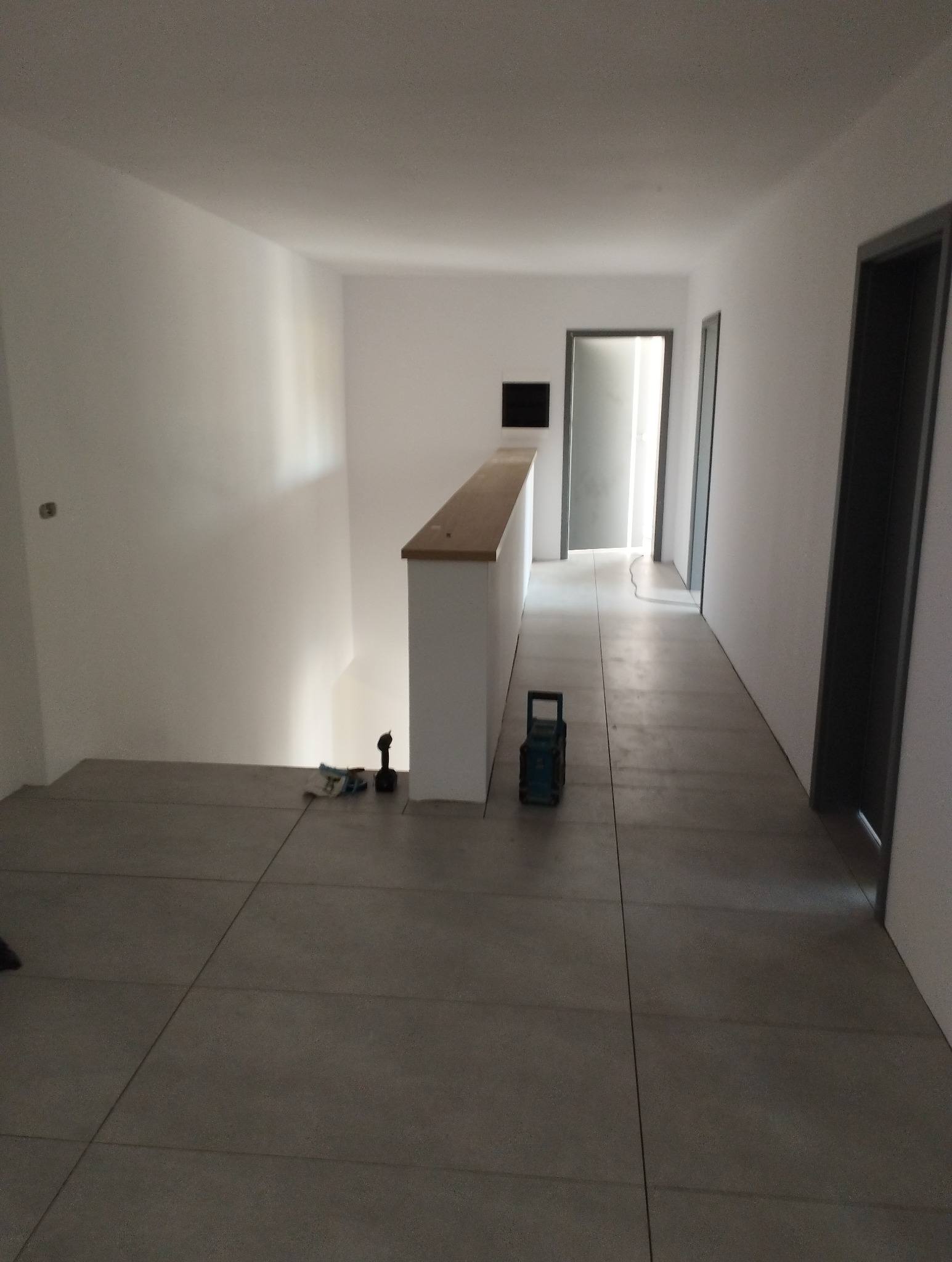 Wnętrze domu w trakcie wykańczania: szara podłoga z płytek, białe ściany, drewniana balustrada, widoczne narzędzia (wiertarka, lampa akumulatorowa) sugerujące trwające prace.