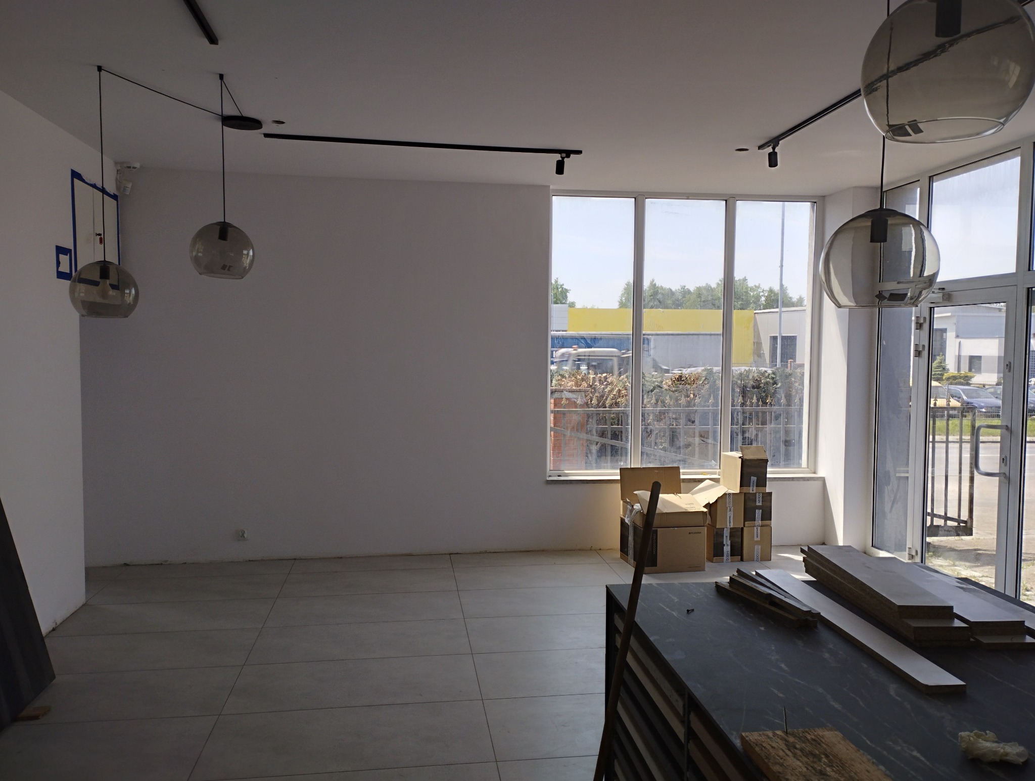 Pusta przestrzeń z nową, jasnoszarą podłogą z płytek, przygotowana do wykończenia; widoczne kartony i panele podłogowe, duże okna z widokiem na zewnątrz.