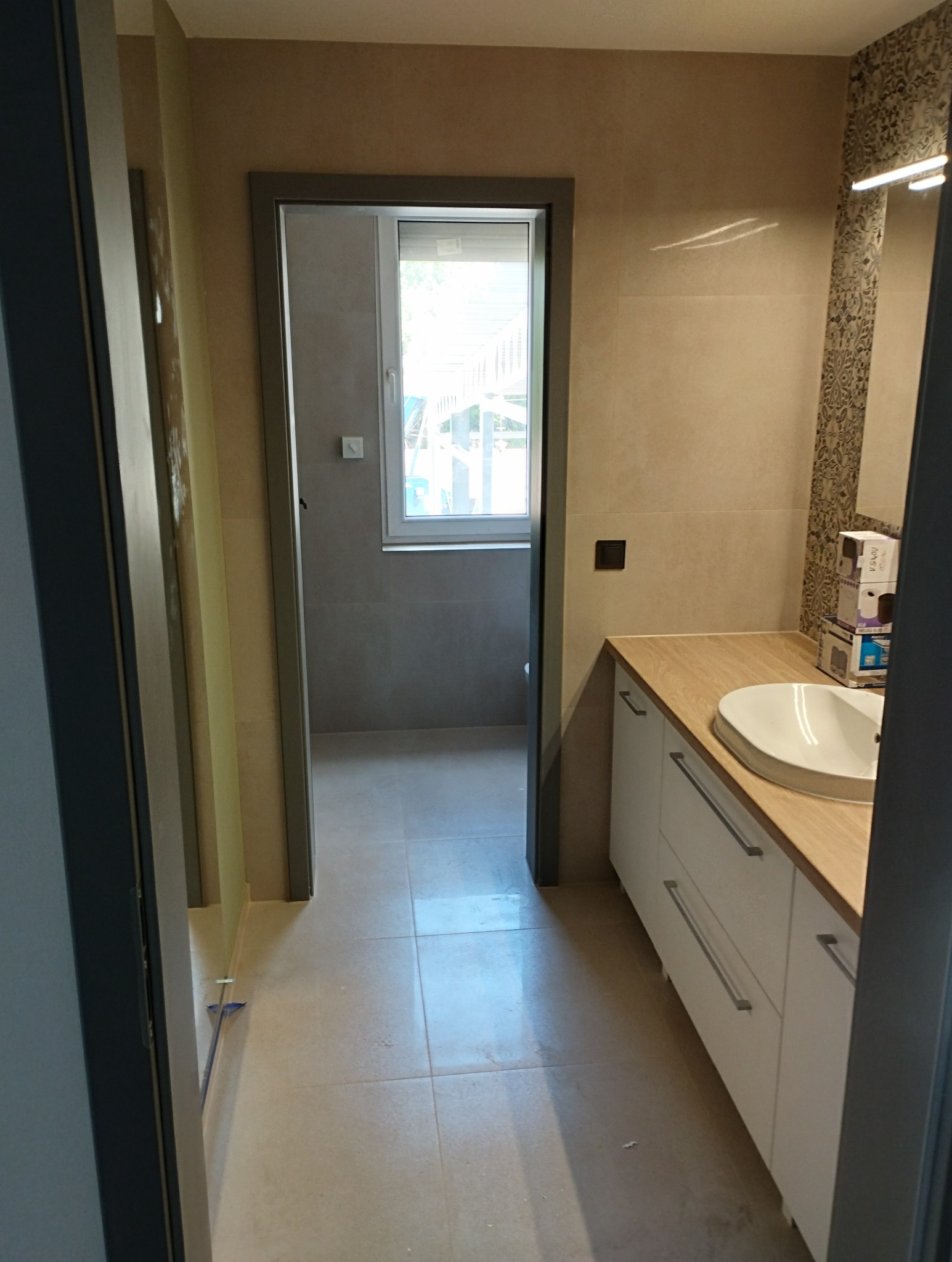 Nowoczesna łazienka z beżowymi płytkami, białymi szafkami pod umywalką z drewnianym blatem i widokiem na okno w drugim pomieszczeniu, uchwycona z perspektywy korytarza podczas remontu.