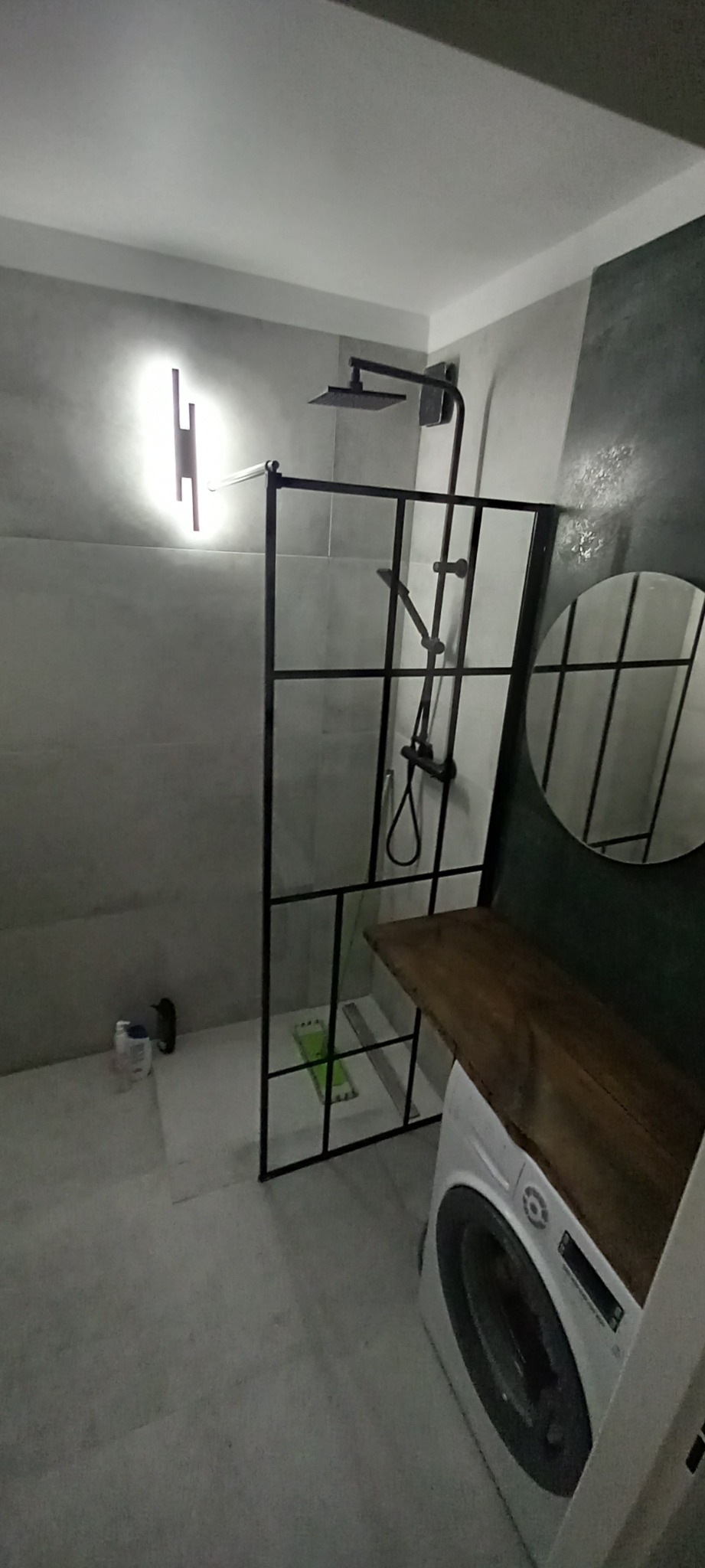 Nowoczesna łazienka z kabiną prysznicową w stylu loft, czarną armaturą, okrągłym lustrem i blatem z drewna nad pralką.