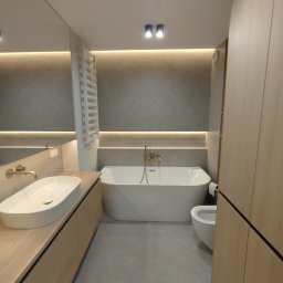 Remonty Łazieki - Nowoczesna łazienka z wanną wolnostojącą, umywalką nablatową i drewnianymi szafkami. Szare płytki i oświetlenie LED tworzą elegancki klimat. Widok z perspektywy wejścia.