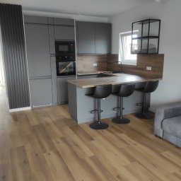 Remonty Łazieki - Nowoczesna kuchnia w szarościach z drewnianym blatem i podłogą. Minimalistyczny design, czarne hokery i sofa w tle. Widok na otwartą przestrzeń.