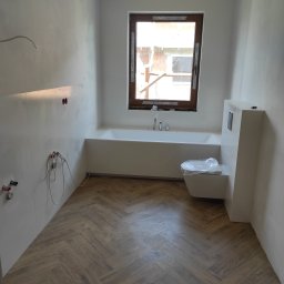 Remonty Łazieki - Jasna łazienka w trakcie wykańczania: białe płytki, drewnopodobna podłoga w jodełkę, wanna pod oknem, wisząca toaleta z folią ochronną. Widoczne kable i niedokończone detale.