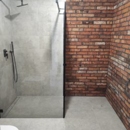Remonty Łazieki - Nowoczesna kabina prysznicowa z ceglaną ścianą w Łodzi. Szklana tafla oddziela strefę mokrą. Minimalistyczny design, szara armatura i płytki.