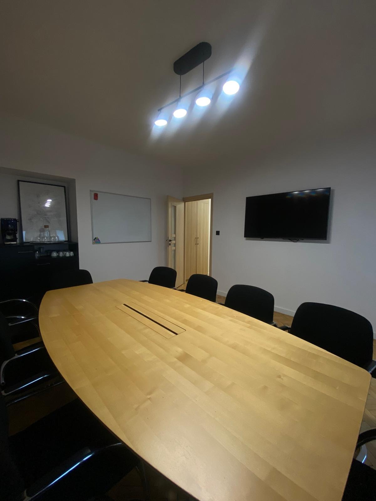 Jasna, drewniana sala konferencyjna z podłużnym stołem, czarnymi krzesłami, białymi ścianami, tablicą suchościeralną i telewizorem, oświetlona lampą z czterema kulistymi żarówkami.