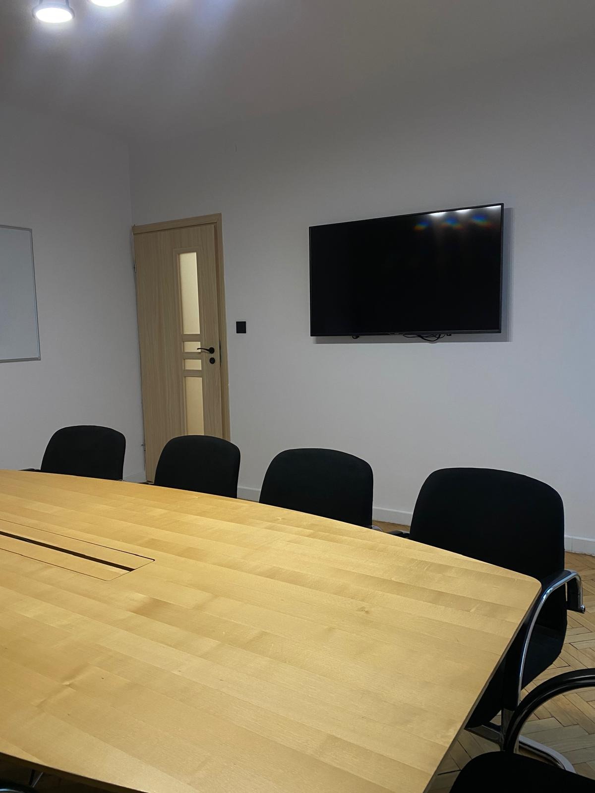 Jasna, drewniana sala konferencyjna z dużym stołem, czarnymi krzesłami i telewizorem LCD na ścianie, oświetlona światłem z sufitu.