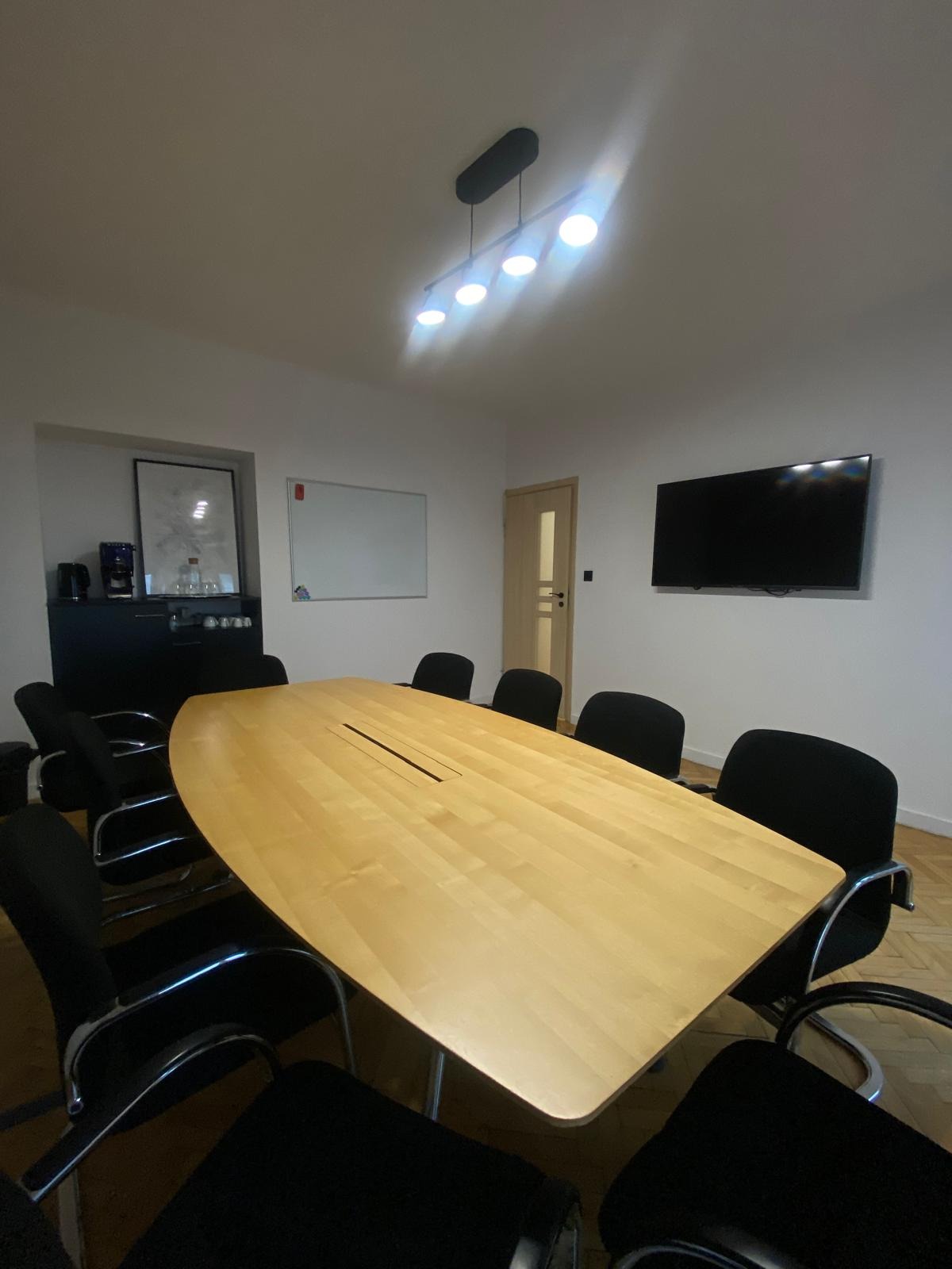 Jasna sala konferencyjna z owalnym stołem z jasnego drewna z czarną listwą na środku, czarne krzesła z metalowymi podłokietnikami, biała tablica, telewizor na ścianie i czarna szafka z ekspresem...