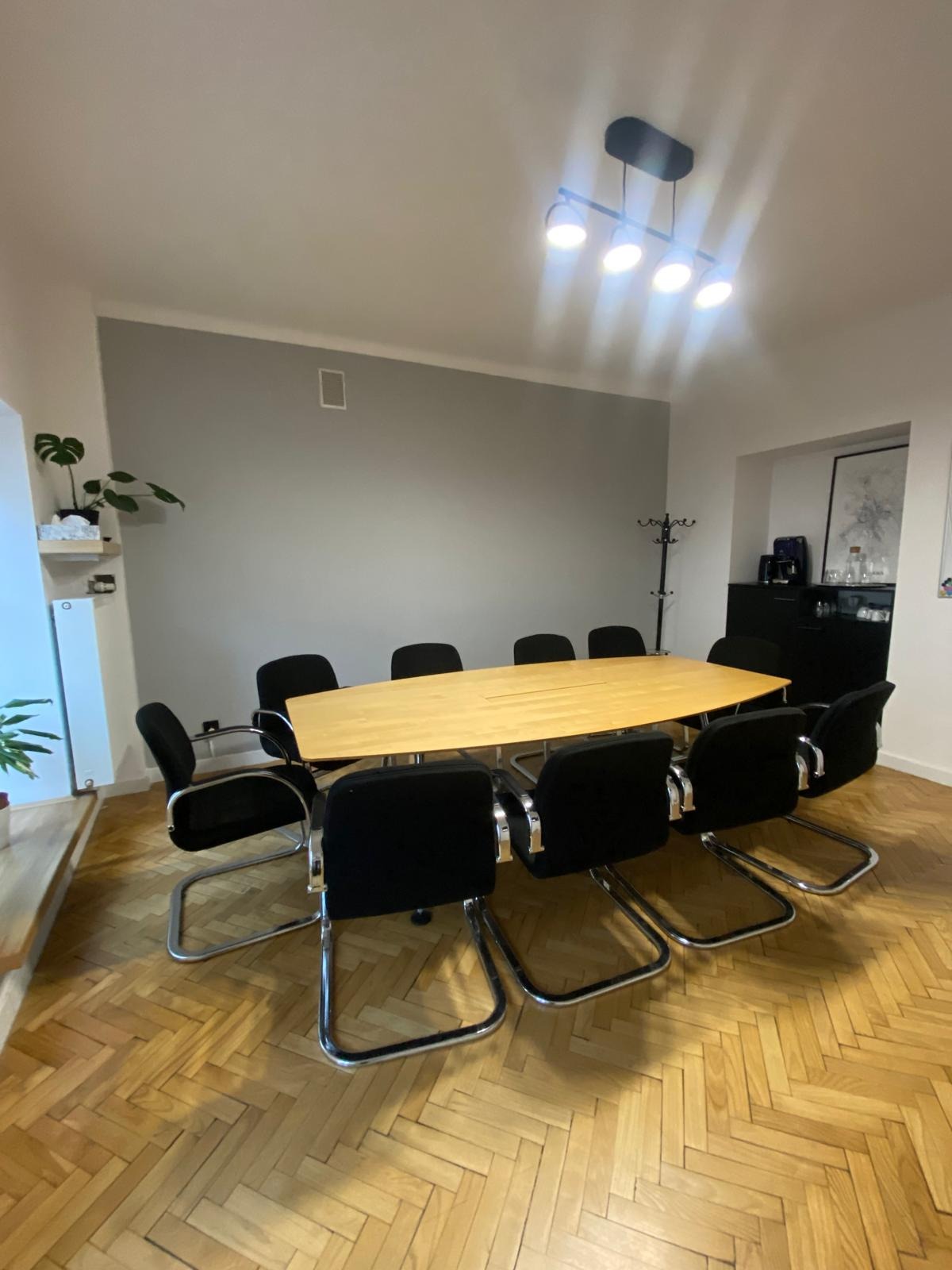 Sala konferencyjna z parkietem w jodełkę, jasnym stołem i czarnymi krzesłami na metalowych płozach, oświetlona czterema punktowymi lampami.