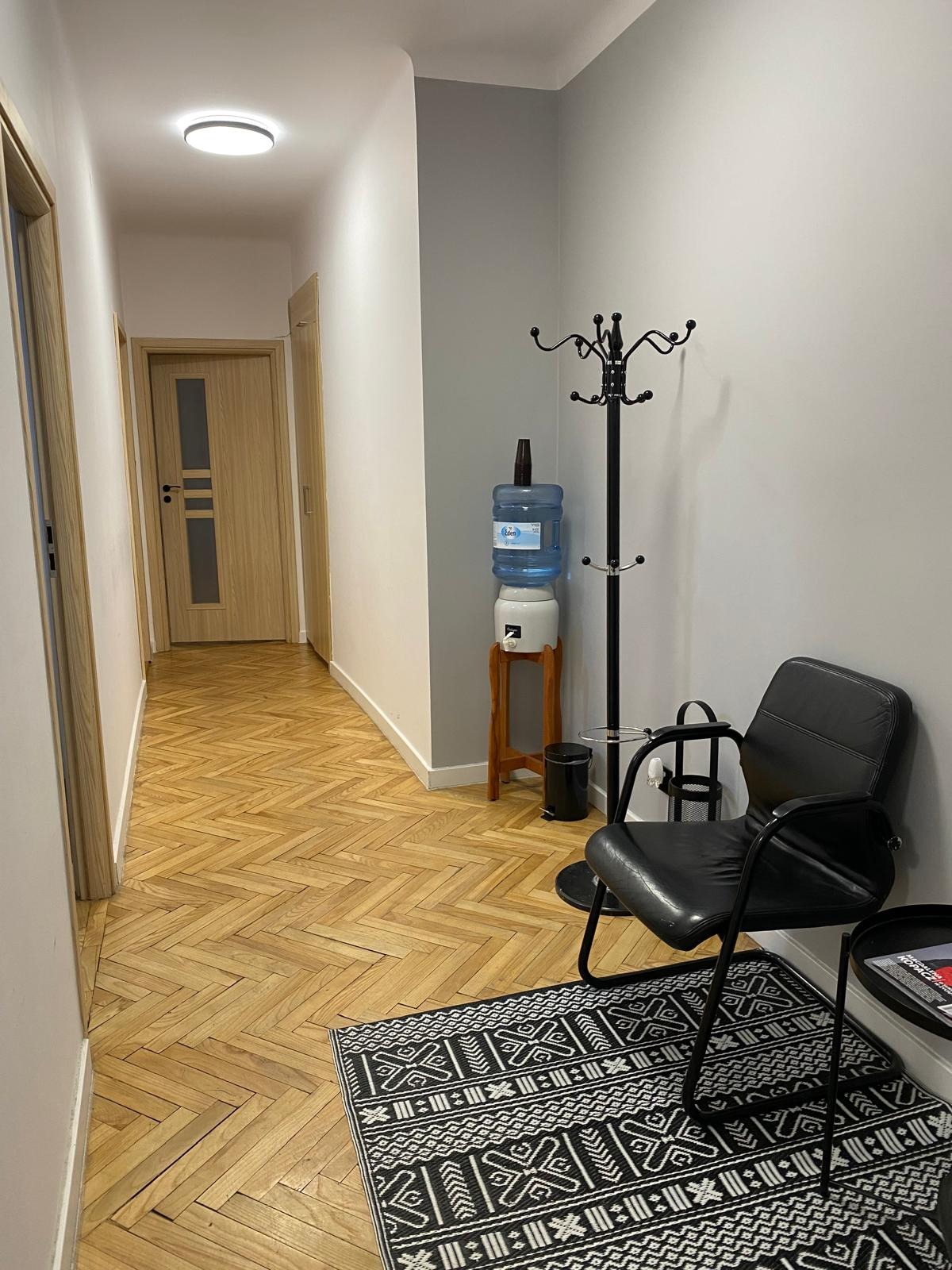 Korytarz biurowy w Warszawie z parkietem w jodełkę, czarnym krzesłem, wieszakiem na ubrania i dystrybutorem wody. W tle widoczne drzwi.