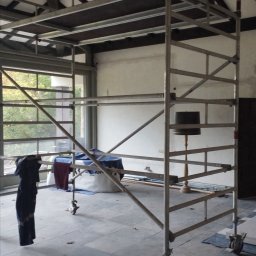 Jerzy Kalinowski - Wnętrze w trakcie prac: metalowe rusztowanie na tle okna i ściany z lampą. Widoczna podłoga z szarych płyt i fragment sufitu z wentylatorem.