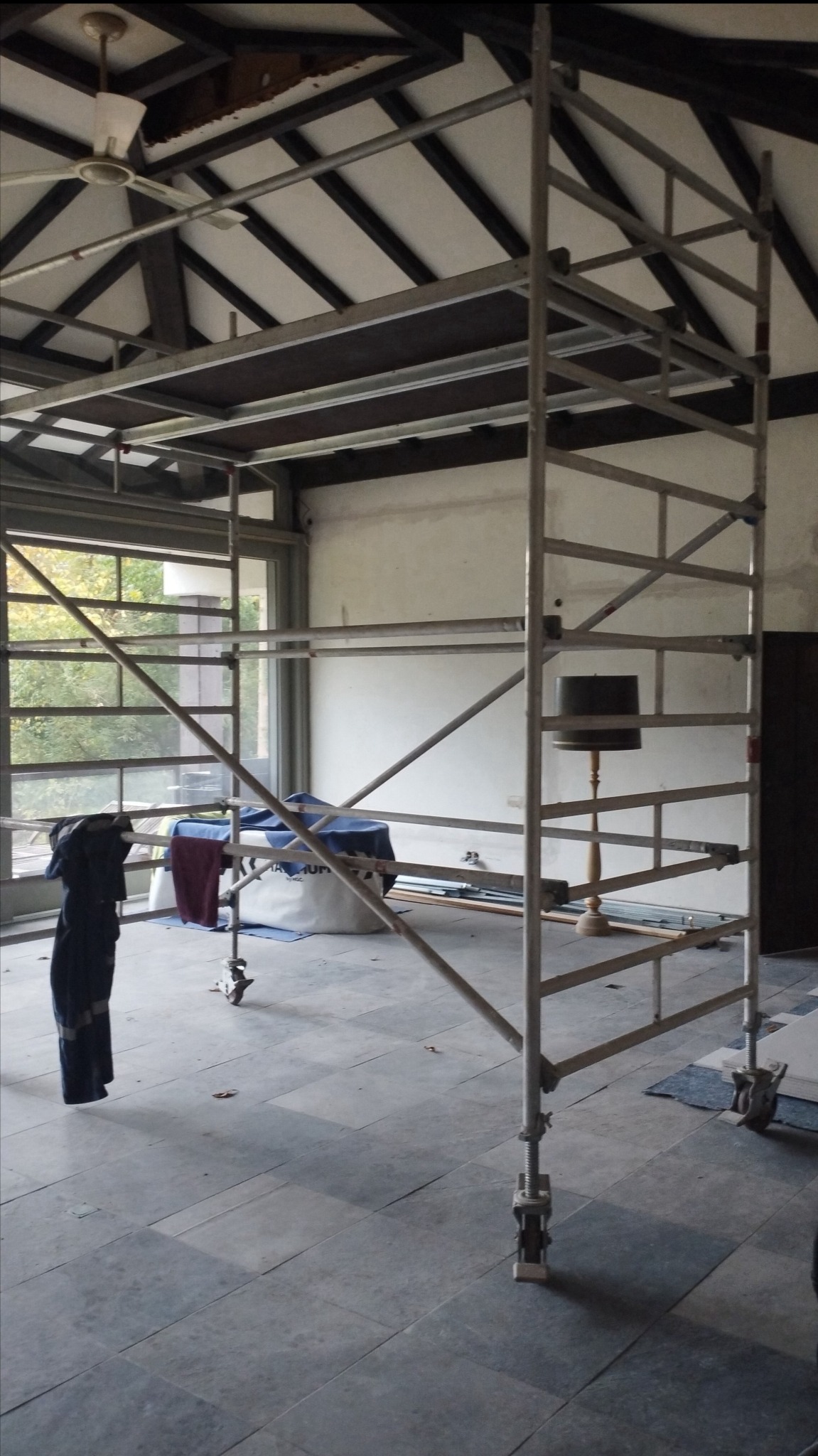 Wnętrze w trakcie prac: metalowe rusztowanie na tle okna i ściany z lampą. Widoczna podłoga z szarych płyt i fragment sufitu z wentylatorem.
