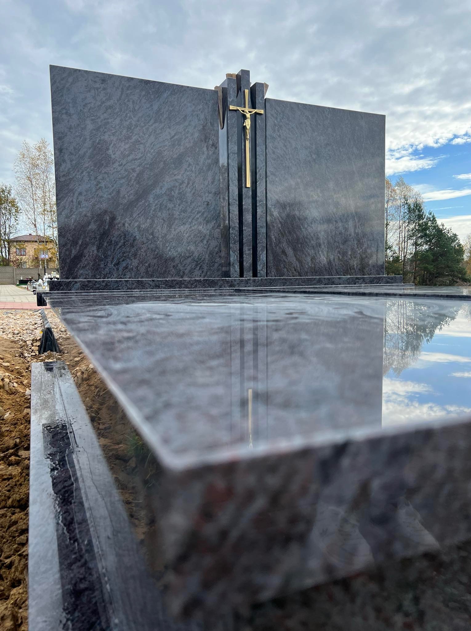Nowoczesny, minimalistyczny nagrobek z ciemnego kamienia z złotym krzyżem, widoczny z perspektywy blatu z odbijającym się niebem.