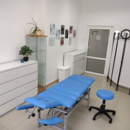 Studio Ruchu i Terapii HOLISTIQ - Jasne wnętrze gabinetu z niebieskim stołem do zabiegów, stołkiem na kółkach, białymi meblami i dekoracjami roślinnymi.