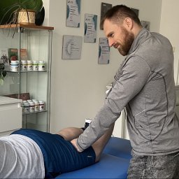 Studio Ruchu i Terapii HOLISTIQ - Mężczyzna w szarej bluzie uciska punkt na udzie pacjenta leżącego na niebieskim stole do masażu; w tle półka z suplementami i dyplomy na ścianie.