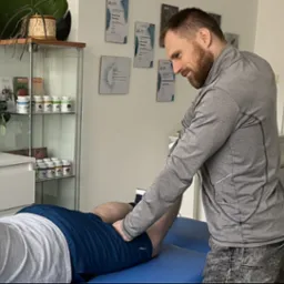 Mężczyzna w szarej bluzie uciska punkt na udzie pacjenta leżącego na niebieskim stole do masażu; w tle półka z suplementami i dyplomy na ścianie.