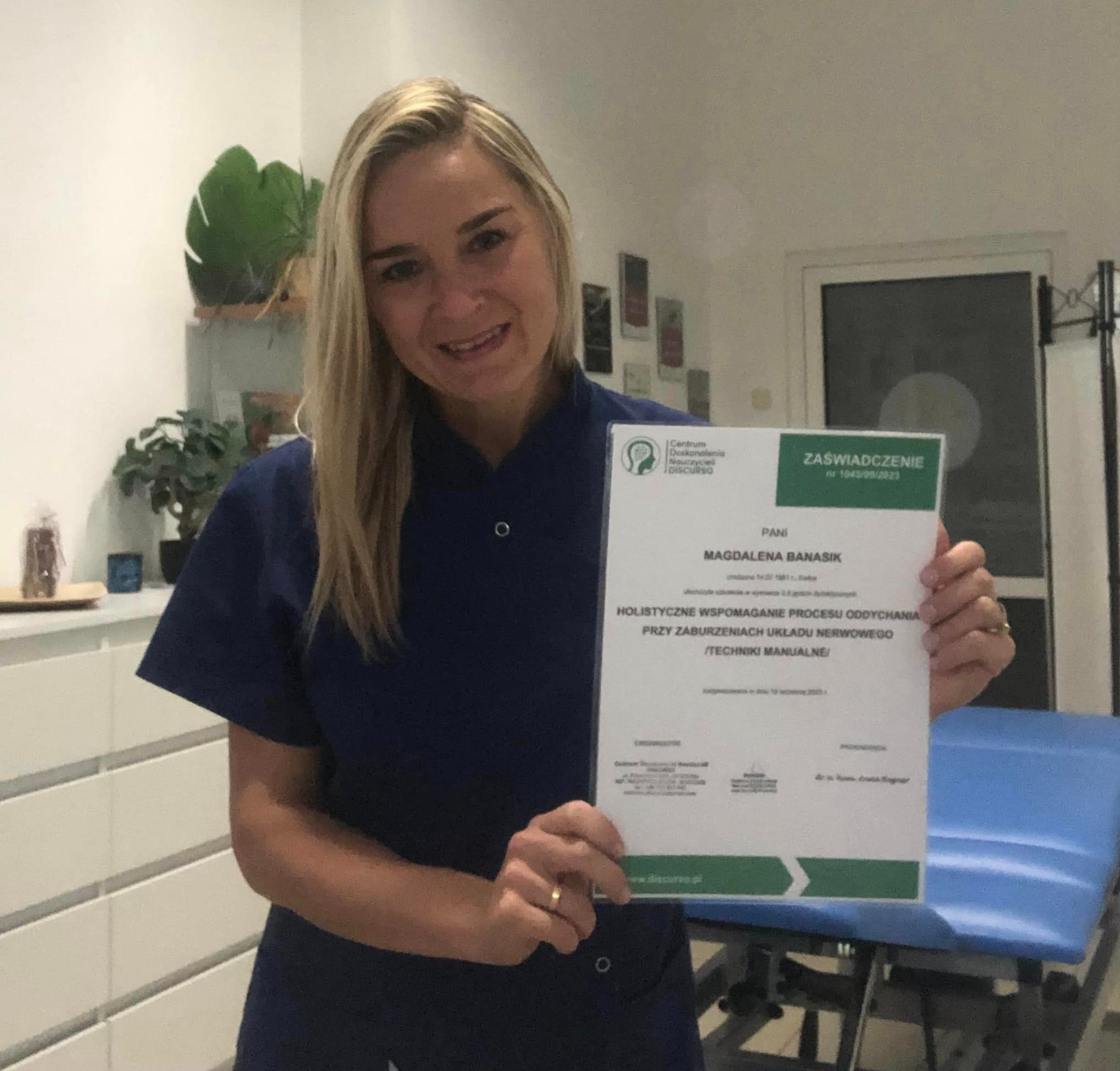 Uśmiechnięta blondynka w granatowym uniformie prezentuje certyfikat ukończenia szkolenia z holistycznego wspomagania procesu oddychania przy zaburzeniach układu nerwowego technikami manualnymi,...