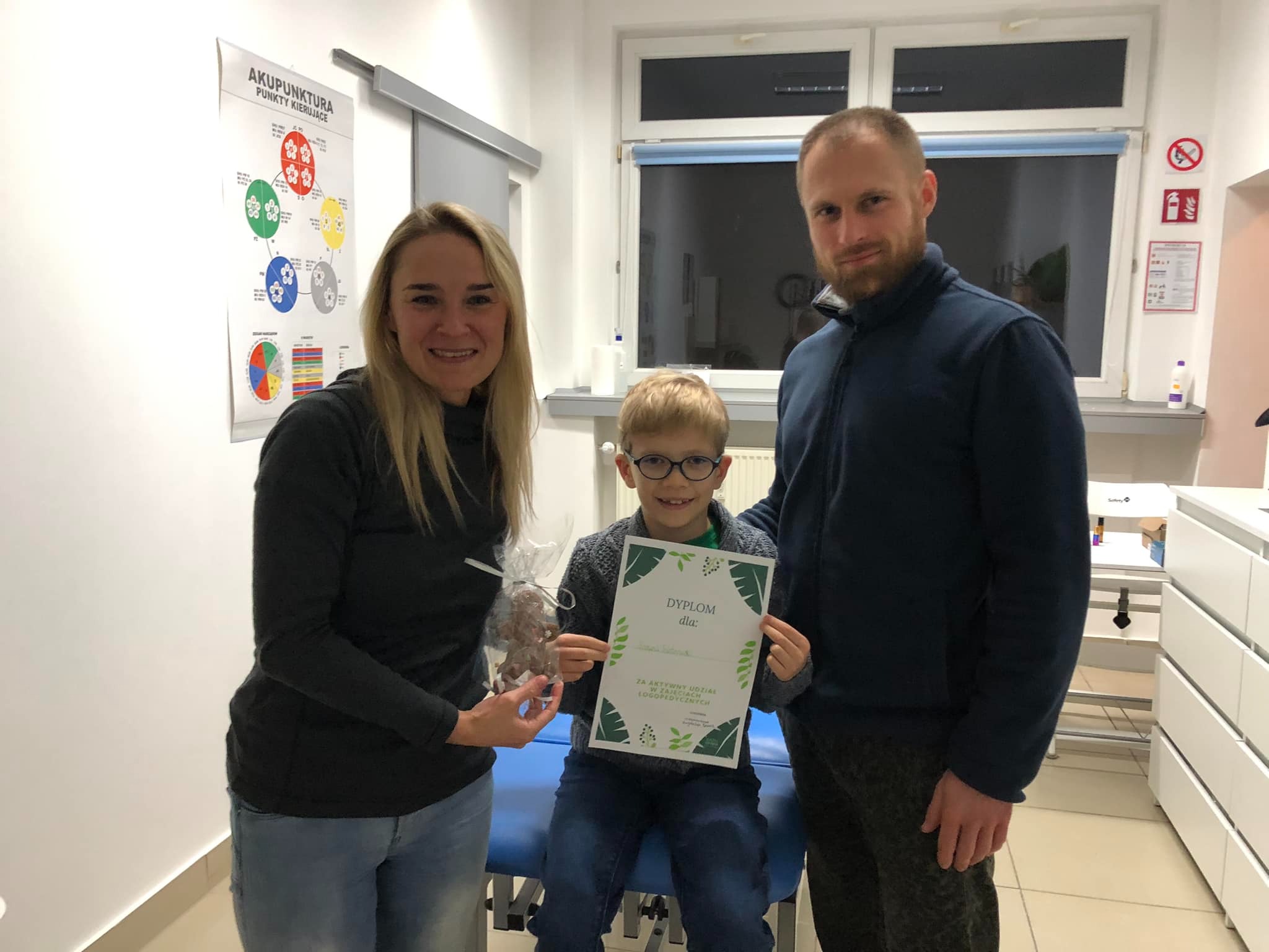Chłopiec w okularach trzyma dyplom za aktywny udział w zajęciach logopedycznych, otoczony przez kobietę z prezentem i mężczyznę, na tle gabinetu z plakatem o akupunkturze.