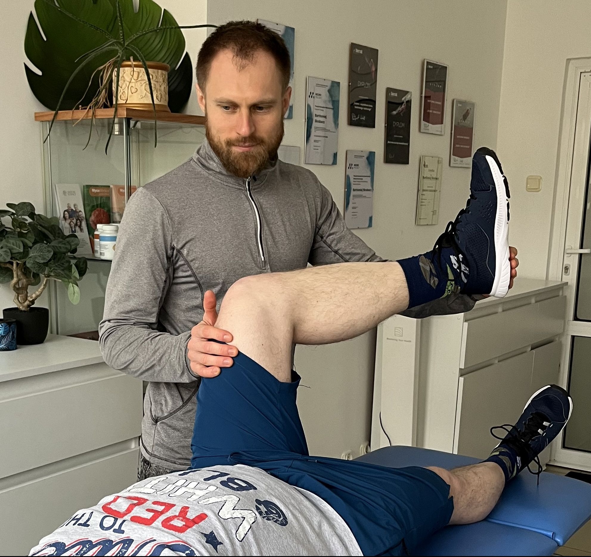 Mężczyzna w szarej bluzie z długim rękawem bada zakres ruchu nogi pacjenta leżącego na leżance, w tle roślina doniczkowa i dyplomy na ścianie.