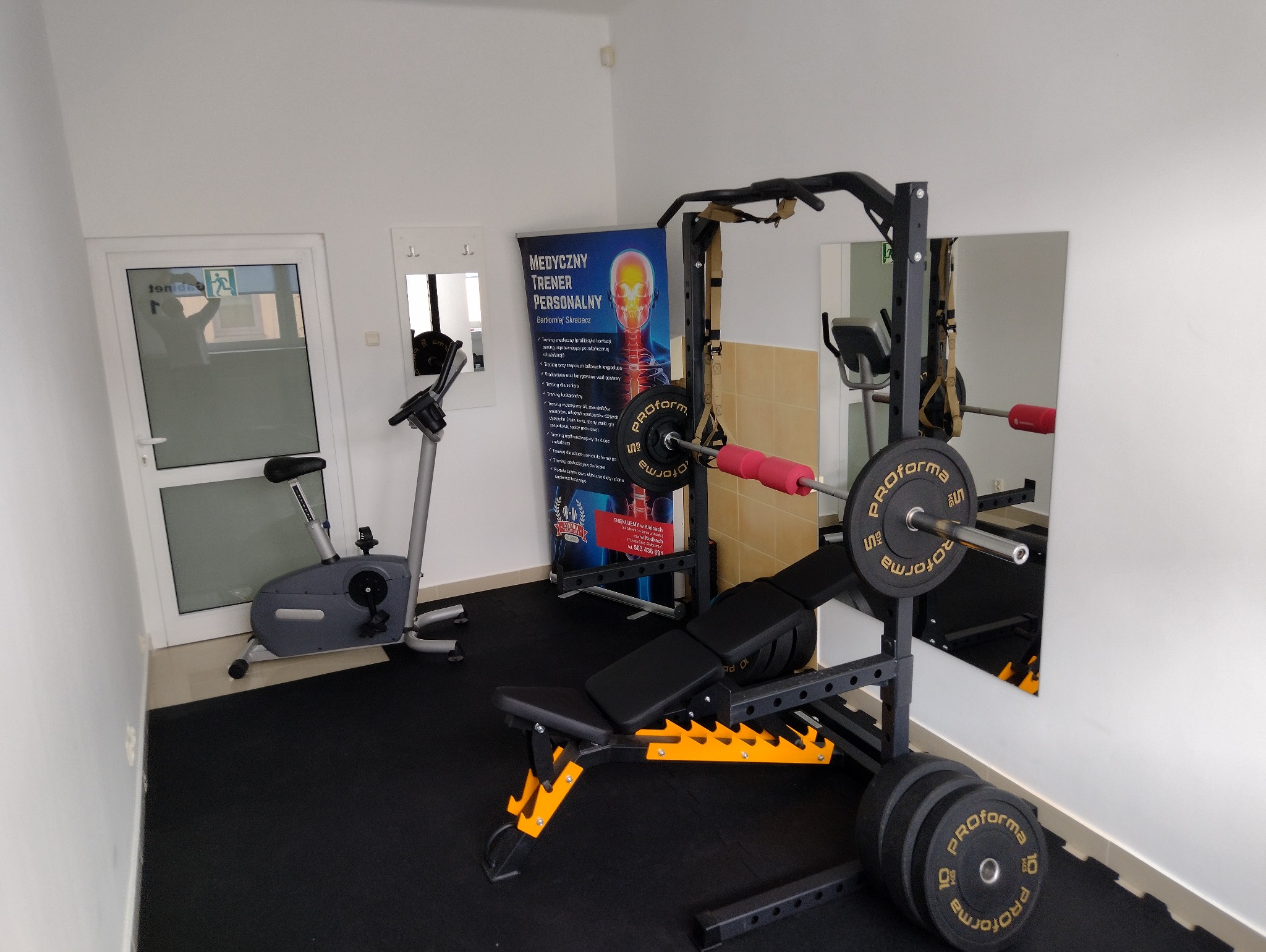 Sprzęt do ćwiczeń w siłowni: ławka regulowana z obciążeniami, rower stacjonarny i plakat z napisem 'Medyczny Trener Personalny'.