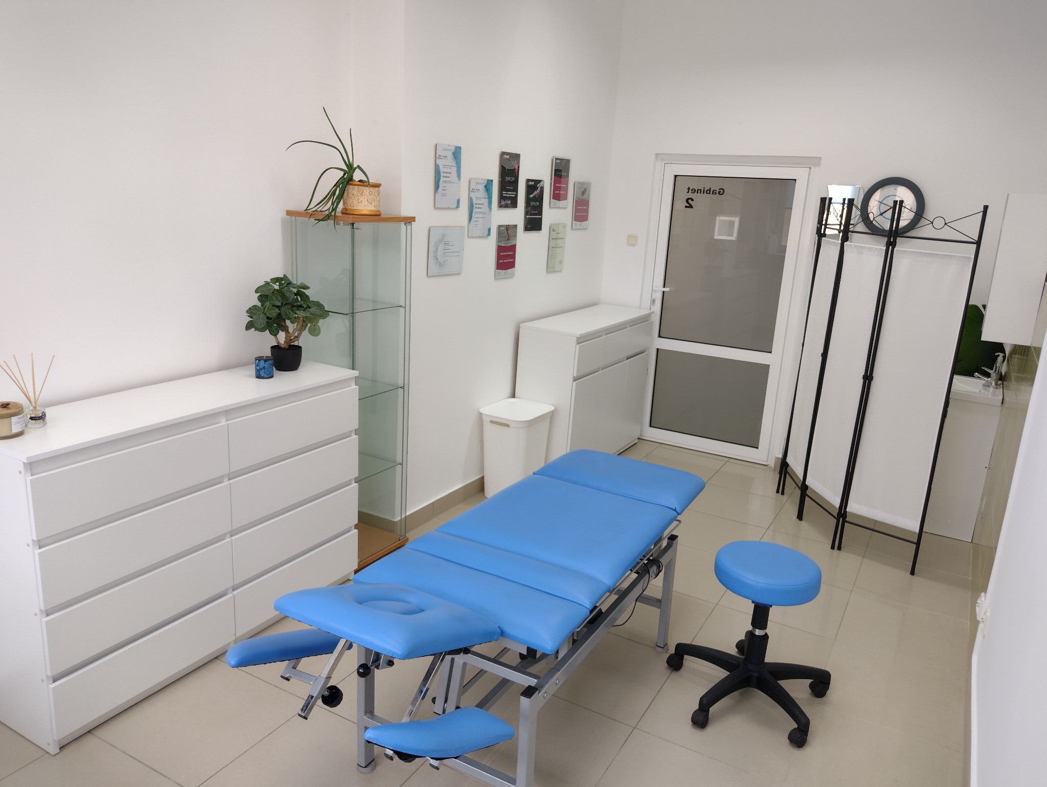 Jasne wnętrze gabinetu z niebieskim stołem do zabiegów, stołkiem na kółkach, białymi meblami i dekoracjami roślinnymi.