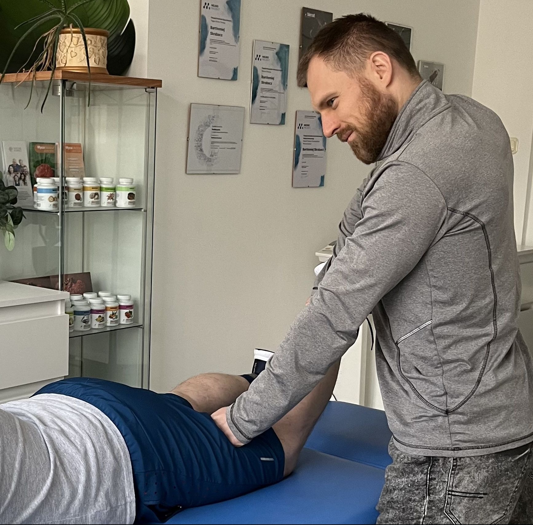 Mężczyzna w szarej bluzie uciska punkt na udzie pacjenta leżącego na niebieskim stole do masażu; w tle półka z suplementami i dyplomy na ścianie.