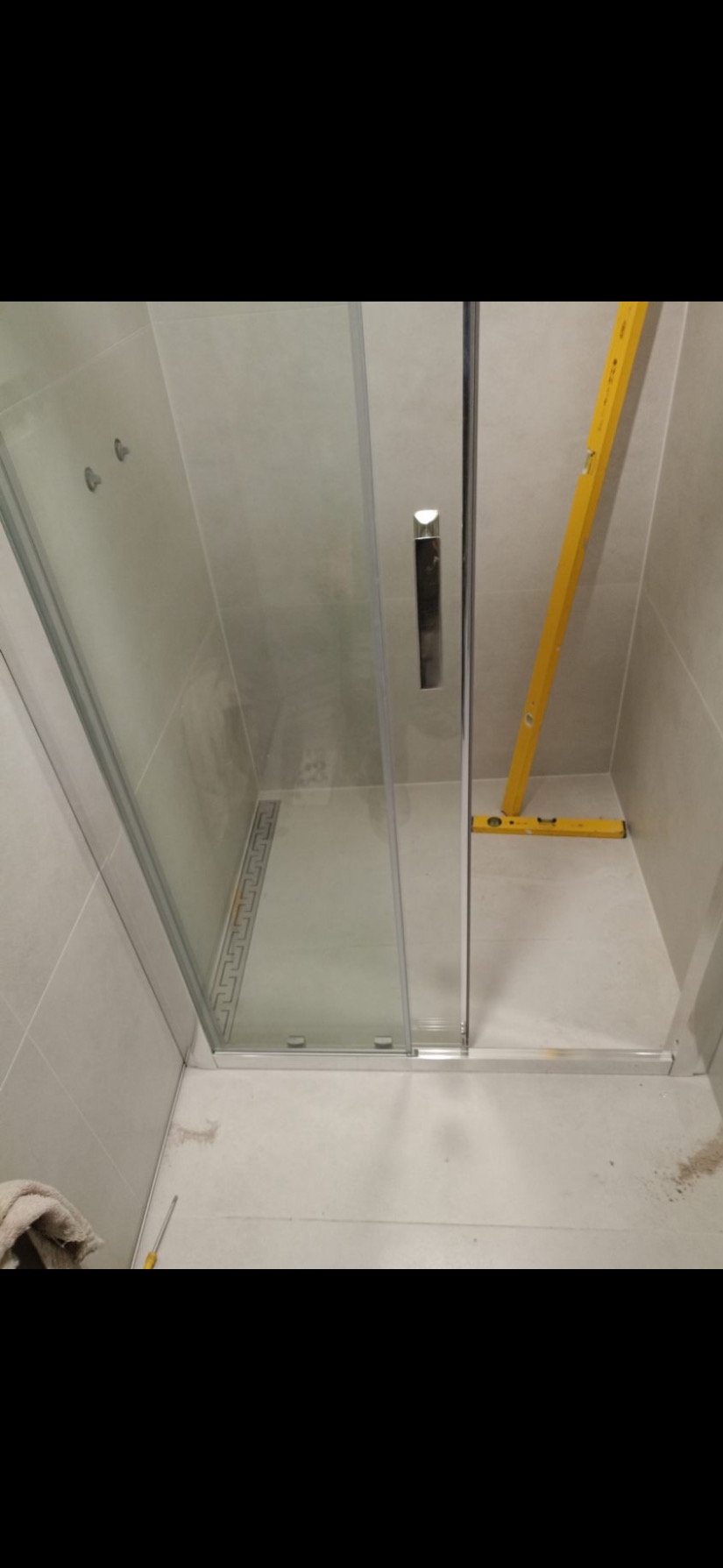 Montaż kabiny prysznicowej: poziomica pionowo oparta o ścianę, mała poziomica na podłodze, widoczny odpływ liniowy z dekoracyjnym wzorem.