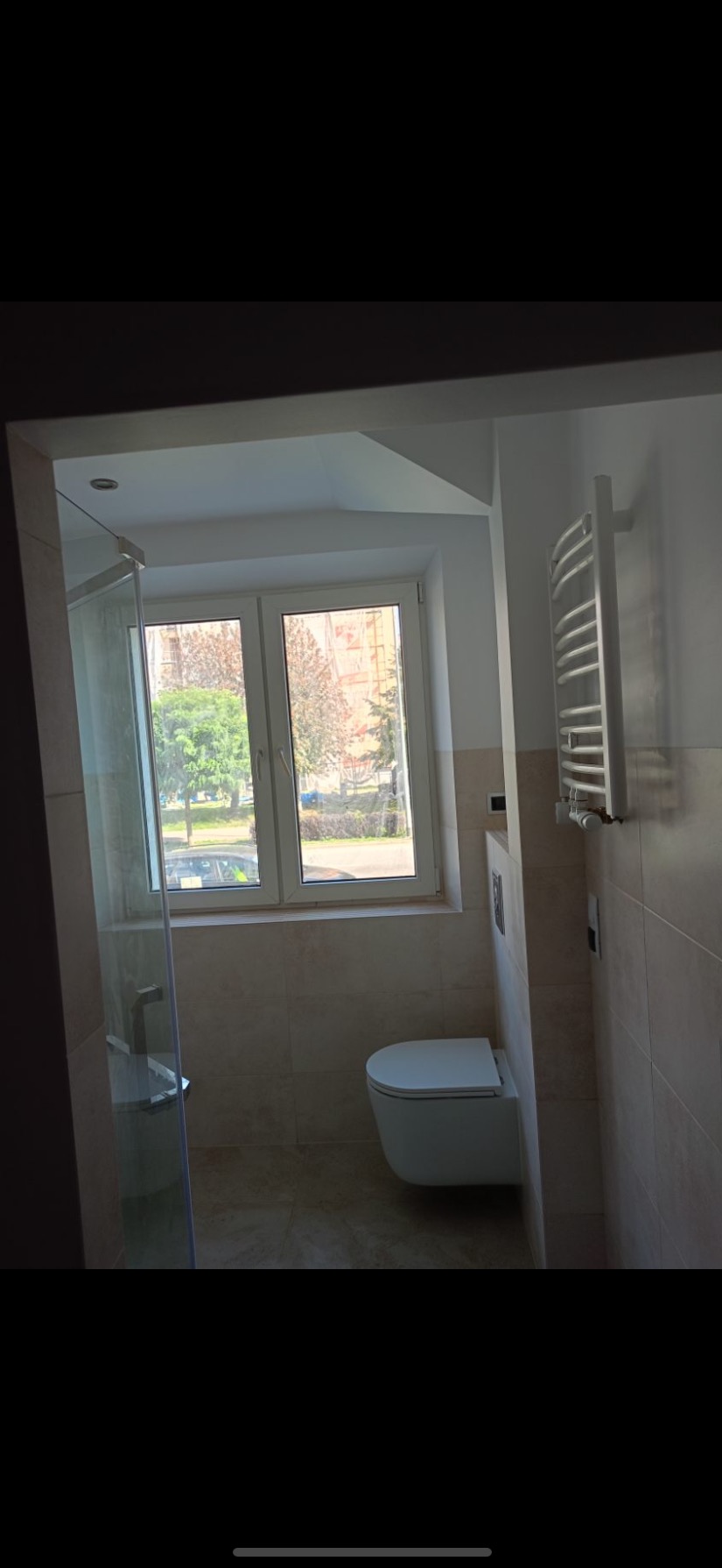 Jasna łazienka z oknem, widok na zewnątrz z zielenią, biała toaleta podwieszana i grzejnik drabinkowy na ścianie pokrytej beżowymi płytkami.