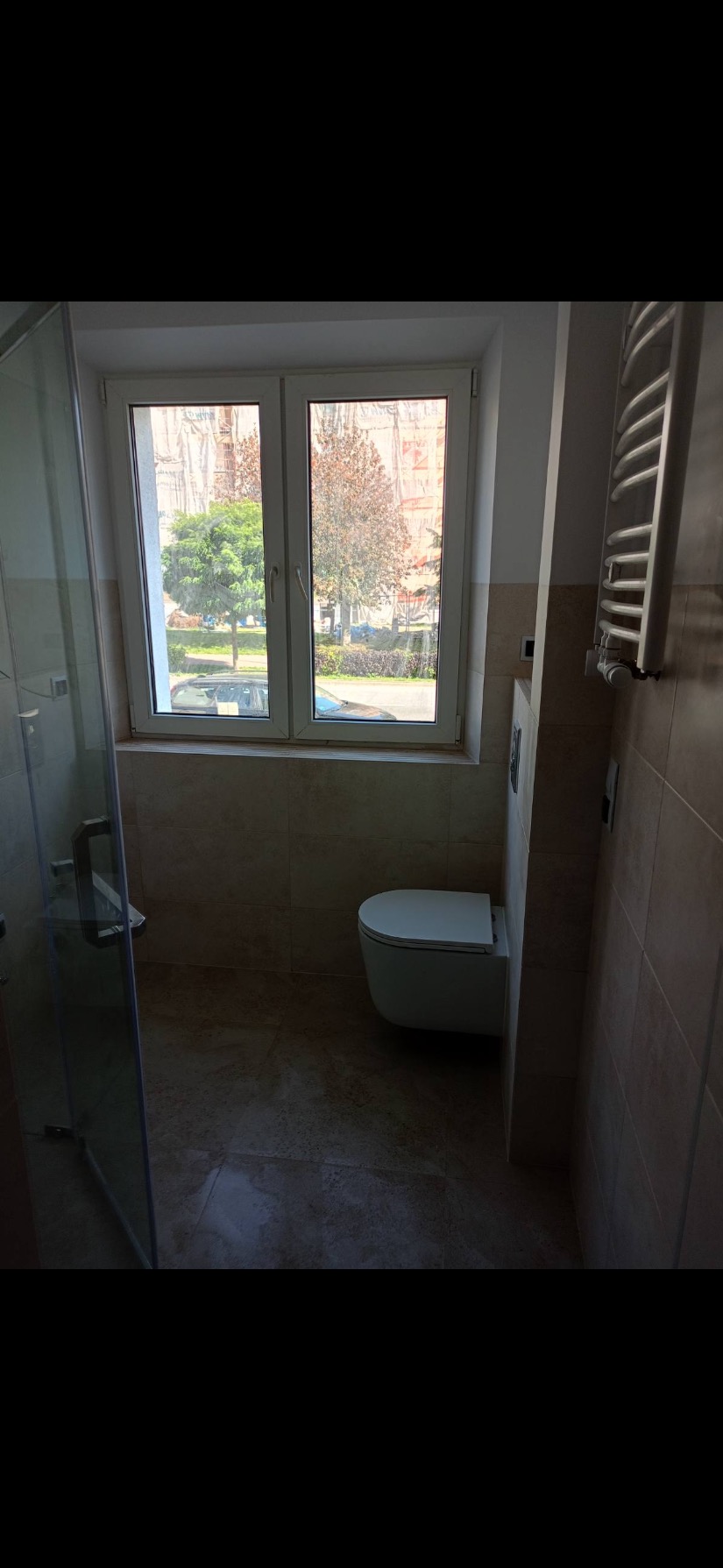 Nowoczesna łazienka z beżowymi płytkami, wiszącą toaletą i szklaną kabiną prysznicową, widok na drzewa za oknem.