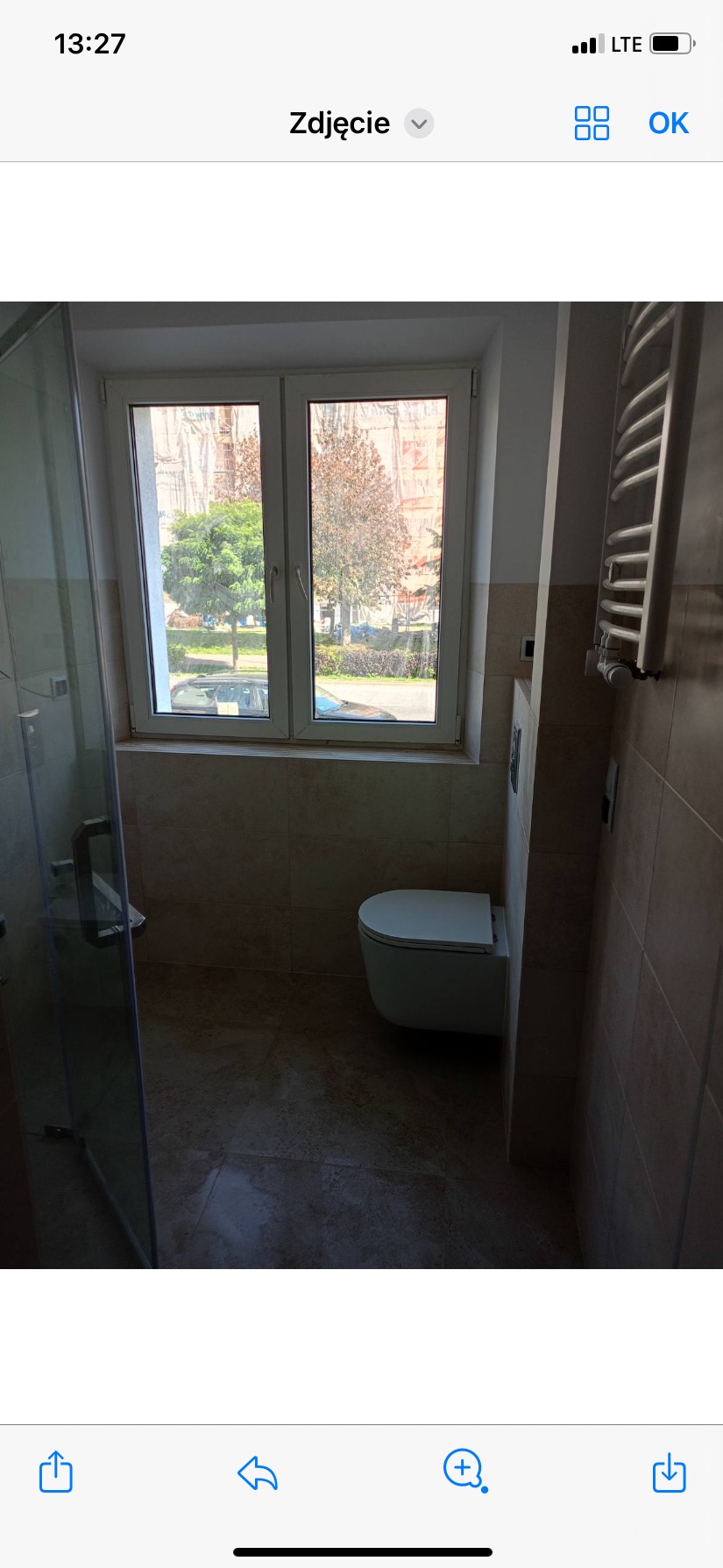 Wykończona łazienka z kabiną prysznicową, podwieszaną toaletą i grzejnikiem drabinkowym, widok na drzewa za oknem.